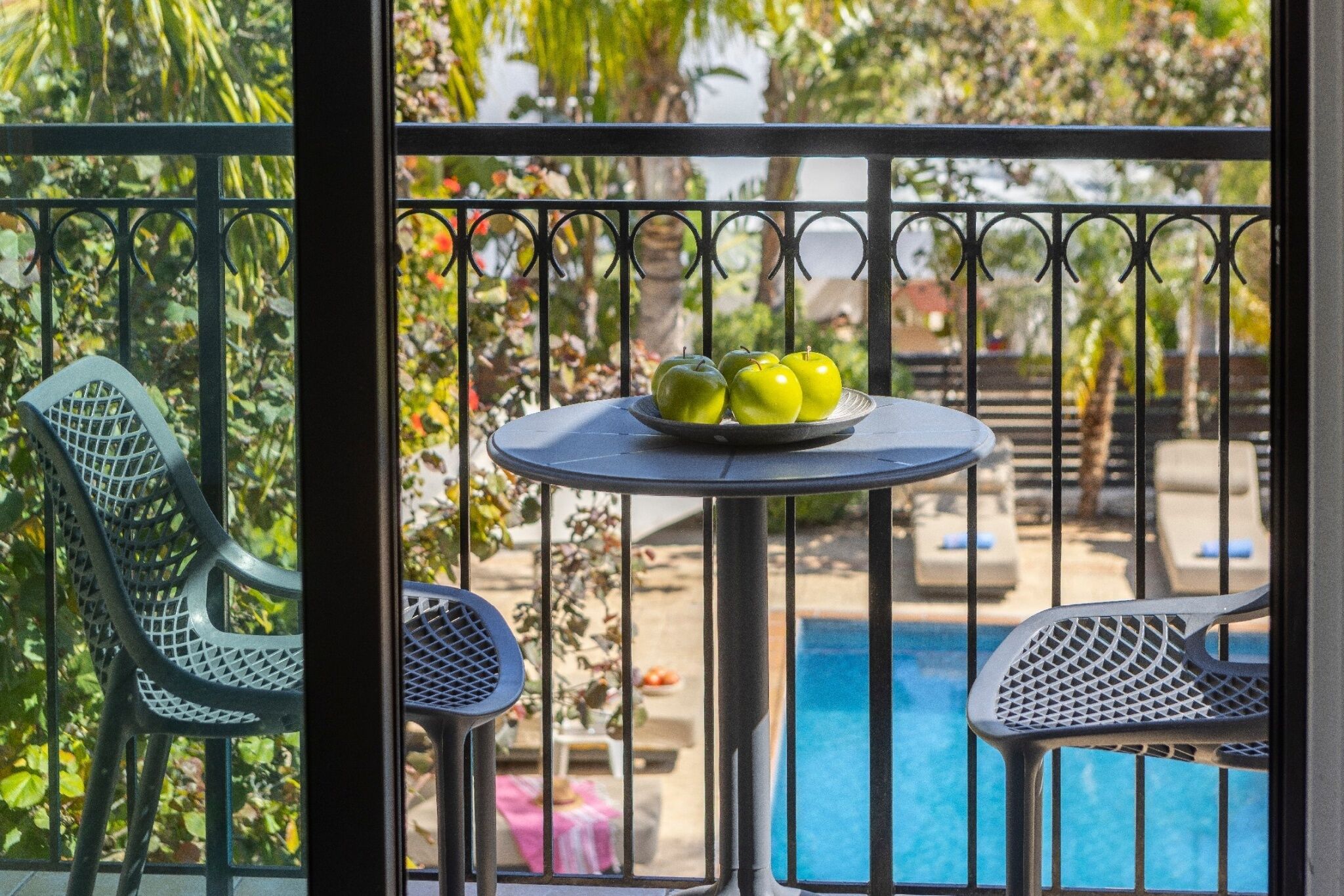 Un balcon avec une table sur laquelle reposent trois pommes, entouré de chaises en métal et avec une piscine en arrière-plan.