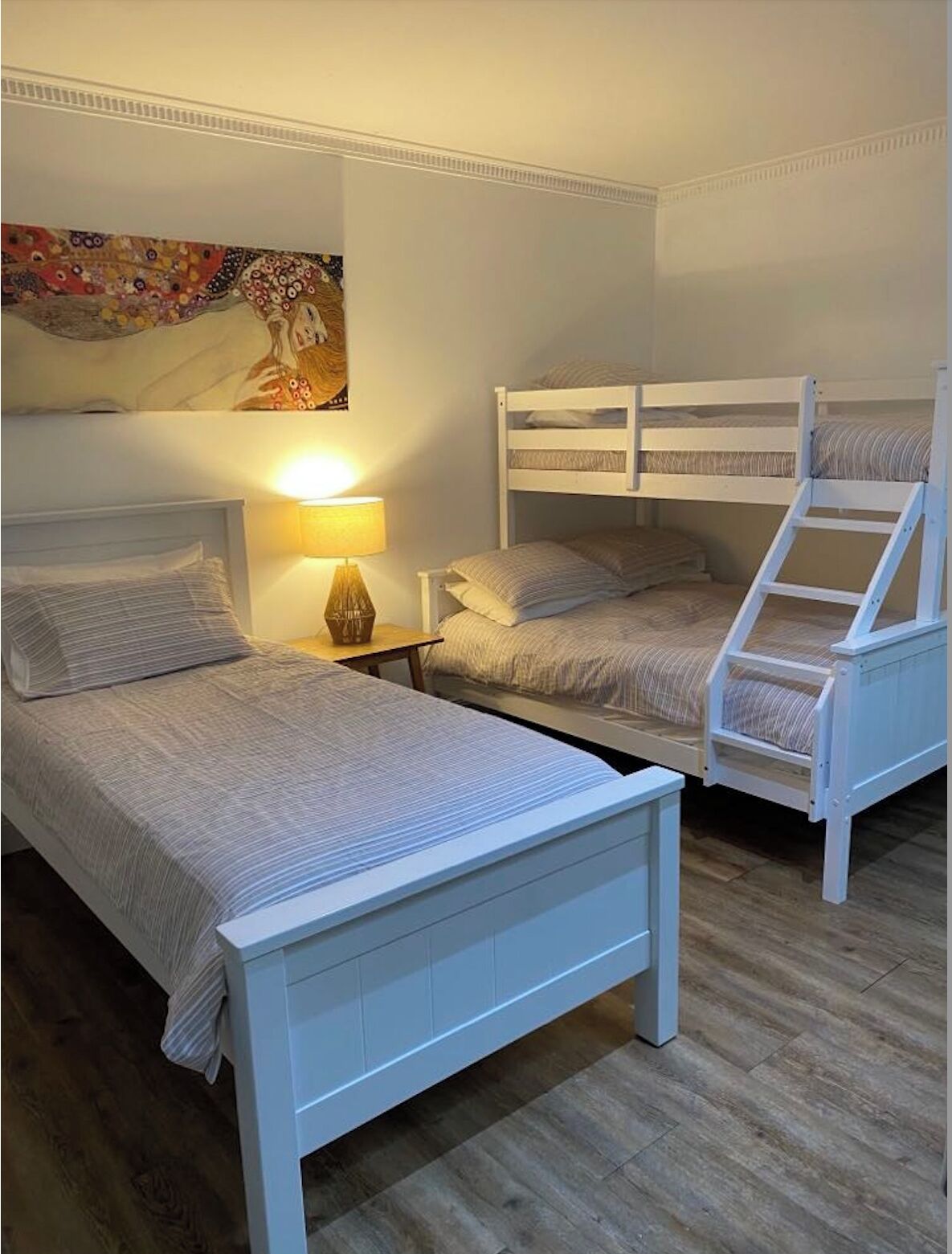 Un dormitorio con dos camas individuales y una litera, una mesita de noche con lámpara y un cuadro en la pared.
