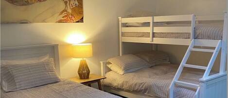 Un dormitorio con dos camas individuales y una litera, una mesita de noche con lámpara y un cuadro en la pared.