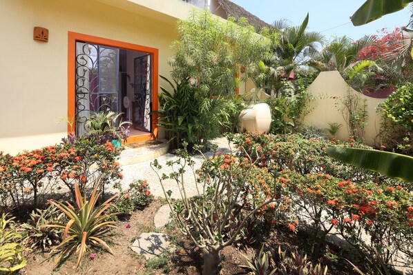 Un jardin avec différentes plantes et fleurs, une entrée d’immeuble dotée d’un cadre orange et un grand objet blanc en arrière-plan.