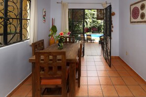 A dining area with a wooden table and chairs, a vase with flowers, and a view of a swimming pool through an open door.