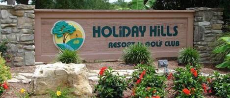 A sign for Holiday Hills Resort & Golf Club with a logo featuring a tree and landscape.