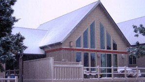 A wooden cabin with a snow-covered roof, a porch with a railing, and a thatched roof structure in the background. - Luxurious 3BR/2BA Eagle Crest Chalet on Golf Course w/ Private Hot Tub & Garage (Redmond)