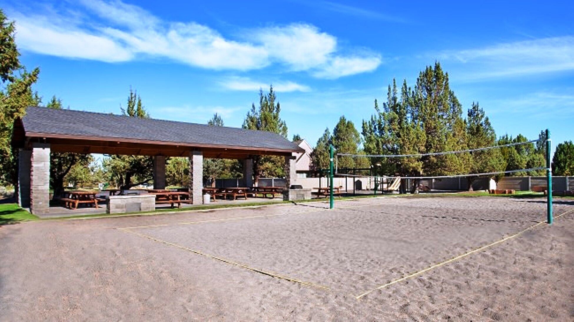A sandy volleyball court with a pavilion, picnic tables, and trees in the background.