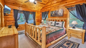 A log cabin bedroom with a wooden bed, a nightstand, a desk, and a window with curtains. - Best Price!!! Cozy Log Cabin — 3 Min to PF Strip, Sleeps 7 + Hot Tub (Pigeon Forge)