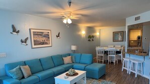 Una sala de estar con sofá modular, una mesa de centro, una zona de comedor con mesa y sillas, y una cocina con mostrador y taburetes.