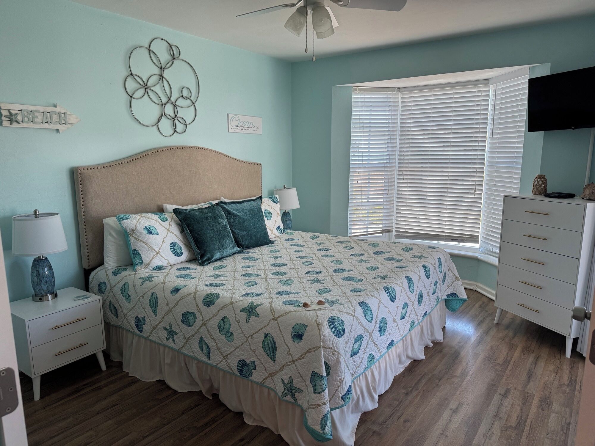 A bedroom with a bed, nightstands, a dresser, a ceiling fan, a window with blinds, and wall decorations.