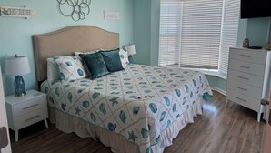 Un dormitorio con cama, mesitas de noche, cómoda, ventilador de techo, ventana con persianas y adornos en la pared.