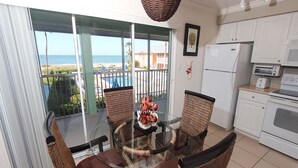 Een eetgedeelte met een glazen tafel en rieten stoelen, een keuken met witte apparatuur, en uitzicht op het strand door een schuifdeur.