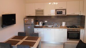 Une cuisine moderne avec des armoires blanches, un four à micro-ondes encastré, une cuisinière et un évier. On y trouve une table à manger avec des chaises, ainsi qu’un téléviseur à écran plat fixé au mur.