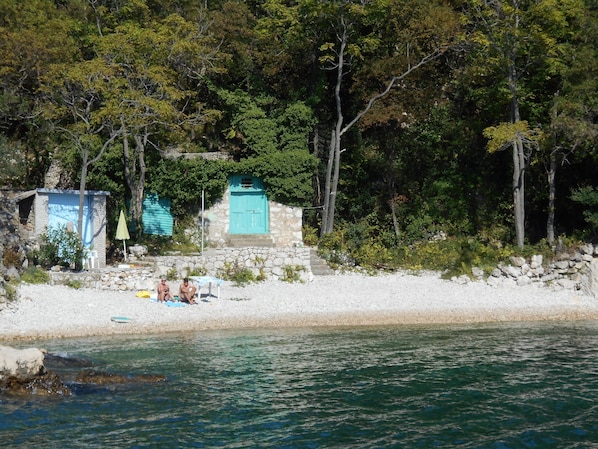 A beach with a stone wall and a small building with a blue door. There are two people sitting on a chair under a blue umbrella.