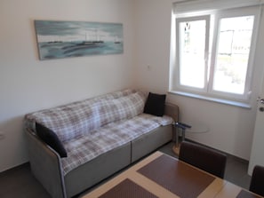 A room with a grey sofa, a painting of a seascape, a window, and a remote control on the sofa.