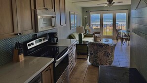 A kitchen with a microwave, stove, and coffee maker. There is a dining area with a table and chairs. The room has a view of the ocean through large windows.