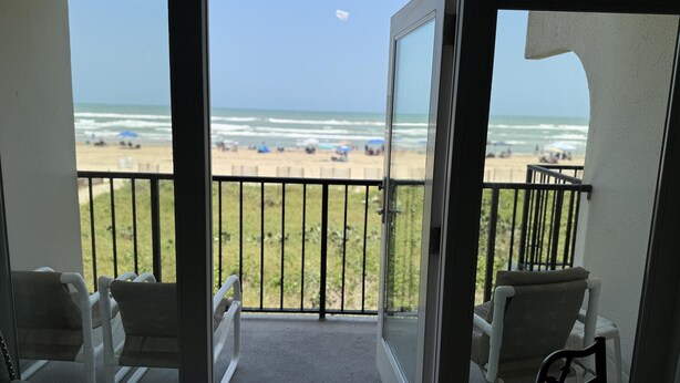 A balcony with a view of the beach, two chairs, and a glass door.