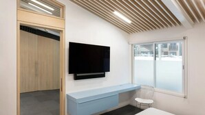 Una habitación moderna con una televisión de pantalla plana montada en la pared, una mesita consola azul claro, una silla blanca y un amplio ventanal con vistas al exterior.