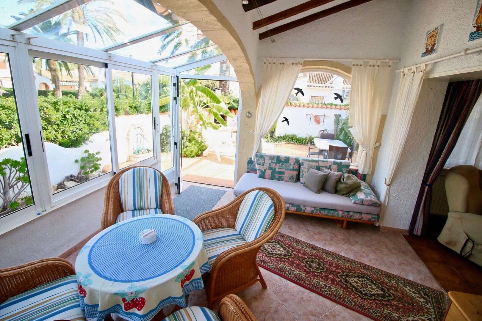 Una habitación iluminada por el sol con un ventanal de vidrio que da a un patio, muebles de mimbre, una alfombra estampada y vistas a zonas verdes y edificios.