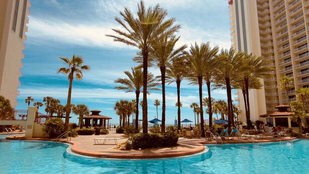 A swimming pool surrounded by palm trees and a high-rise building. - Gulf-Front Corner Unit~Same-Floor Parking~Wrap Balcony~FREE Beach Chairs~Sleep 8 (Panama City Beach)