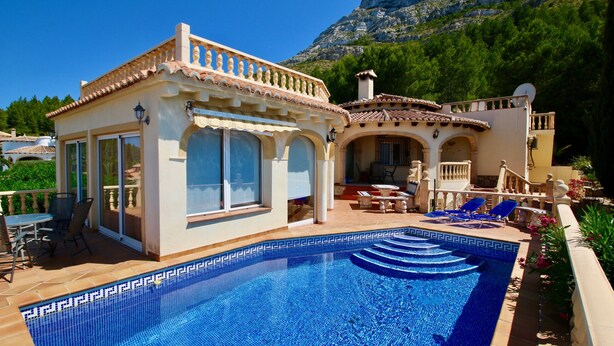 A poolside area with a clear blue pool, outdoor furniture, and a mountain backdrop.