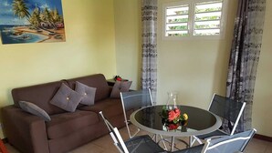 A living room with a brown sofa, grey cushions, a glass coffee table with flowers, and a window with patterned curtains. - Your studio in the Tropics of Martinique. 5 minutes walk from the beach! (Les Trois-Îlets)