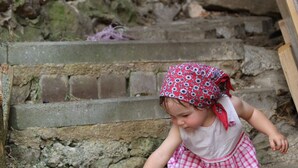 Un niño alimenta a un gato en un entorno rústico al aire libre, con escalones de piedra y detalles de madera.