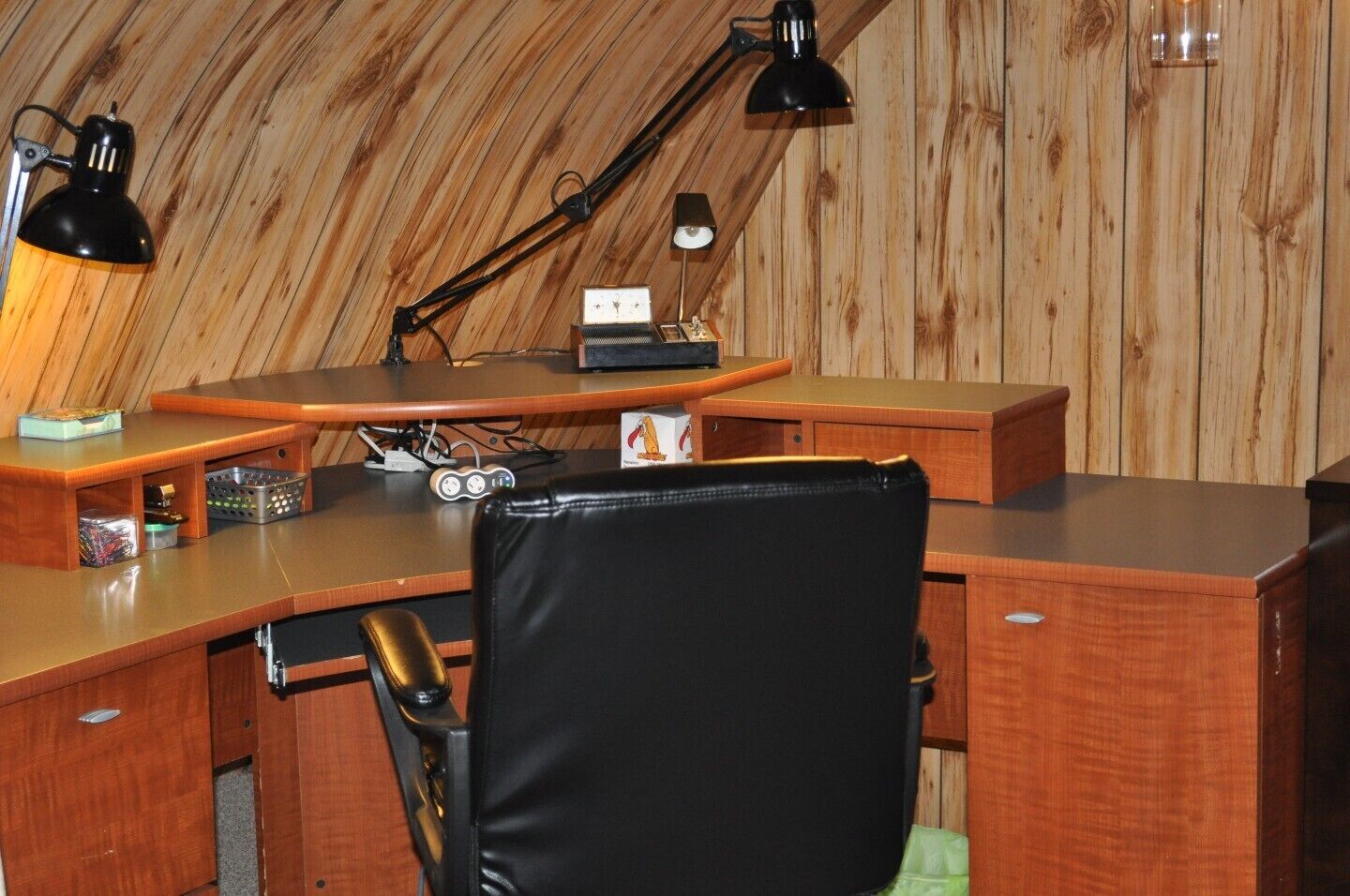 Une pièce aux murs lambrissés, comprenant un bureau, deux lampes de bureau noires, une chaise et une petite étagère sur laquelle sont disposés divers objets.