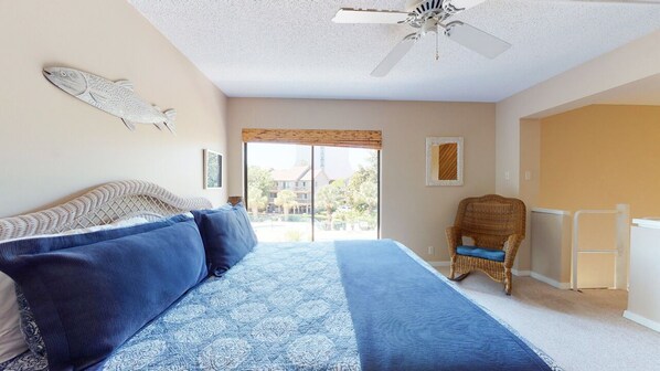 A bedroom with a bed, a ceiling fan, a wicker chair, and a view of the outside.