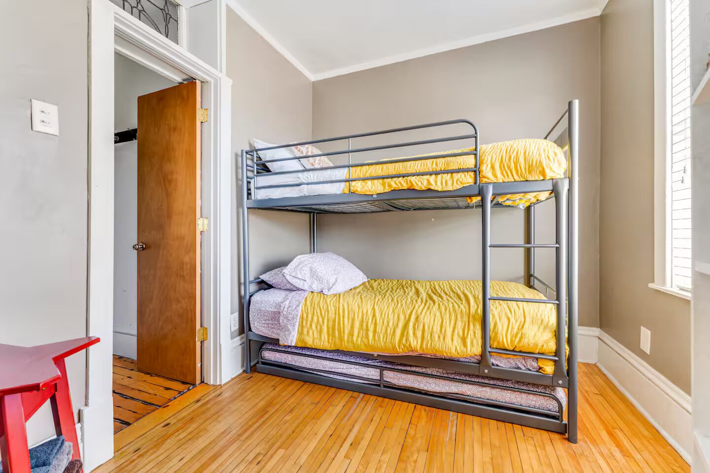 Une chambre avec un lit superposé, un plancher en bois, une chaise rouge et une porte donnant sur une autre pièce.