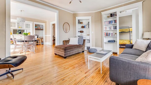 A spacious living area with a wooden floor, a grey sofa, a brown ottoman, and a dining area with a white table and chairs.
