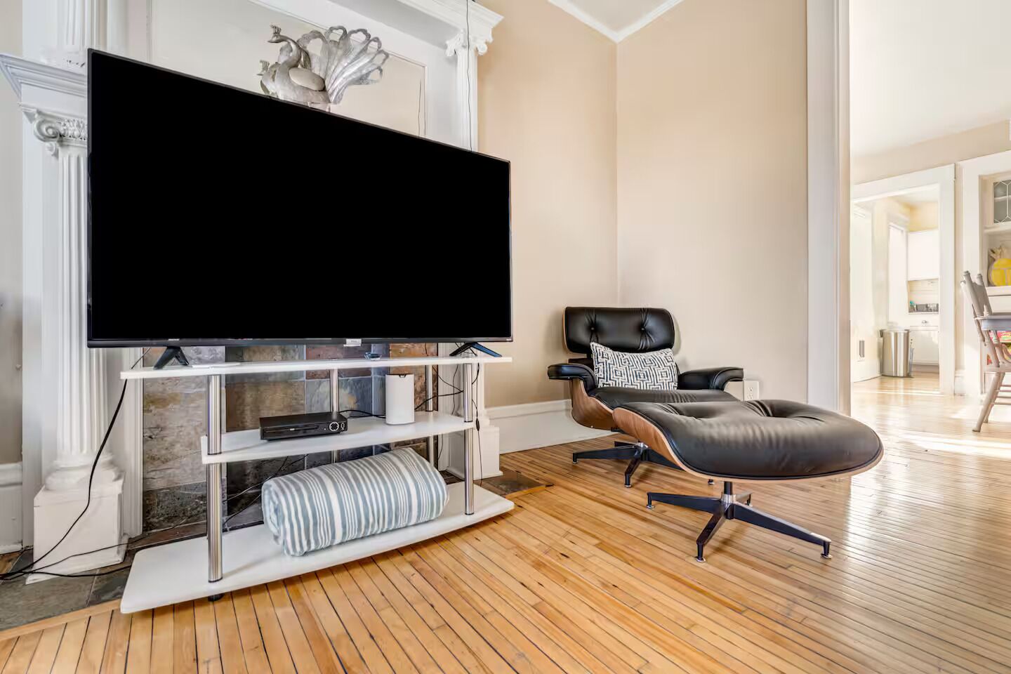 A living room with a large flat-screen TV on a white stand, a leather recliner with a patterned pillow, and a striped ottoman.