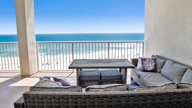 Ein Balkon mit einer Rattan-Couch und einem Tisch, der Blick auf einen Strand mit Menschen und einen strahlend blauen Himmel freigibt.
