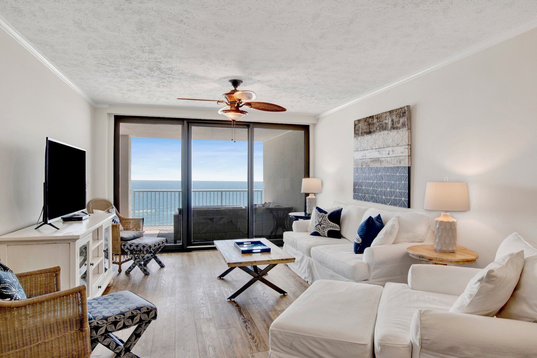 A spacious living room with a large flat-screen TV, a ceiling fan, and a view of the ocean through a sliding glass door.