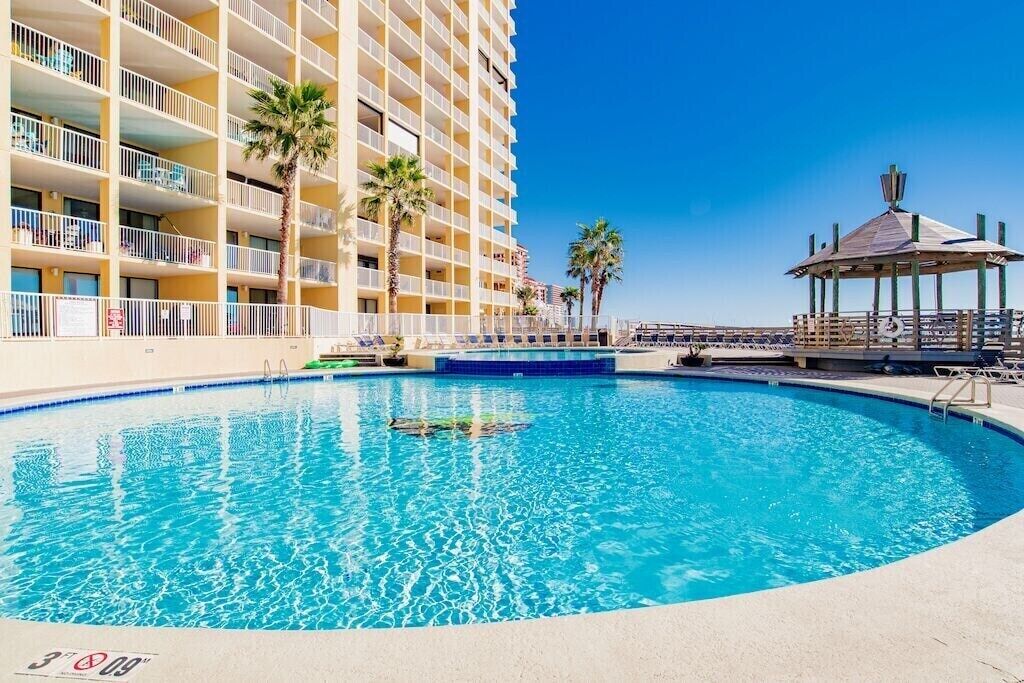 A swimming pool with a clear blue water, surrounded by a building with balconies and palm trees.