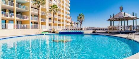 A swimming pool with a clear blue water, surrounded by a building with balconies and palm trees.