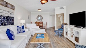Ein modernes Wohnzimmer mit Deckenventilator, weißen Sofas mit blauen Kissen in Musteroptik, einem Holztisch und einem Fernseher auf einem weißen Schrank. An den Wänden befinden sich Kunstwerke.