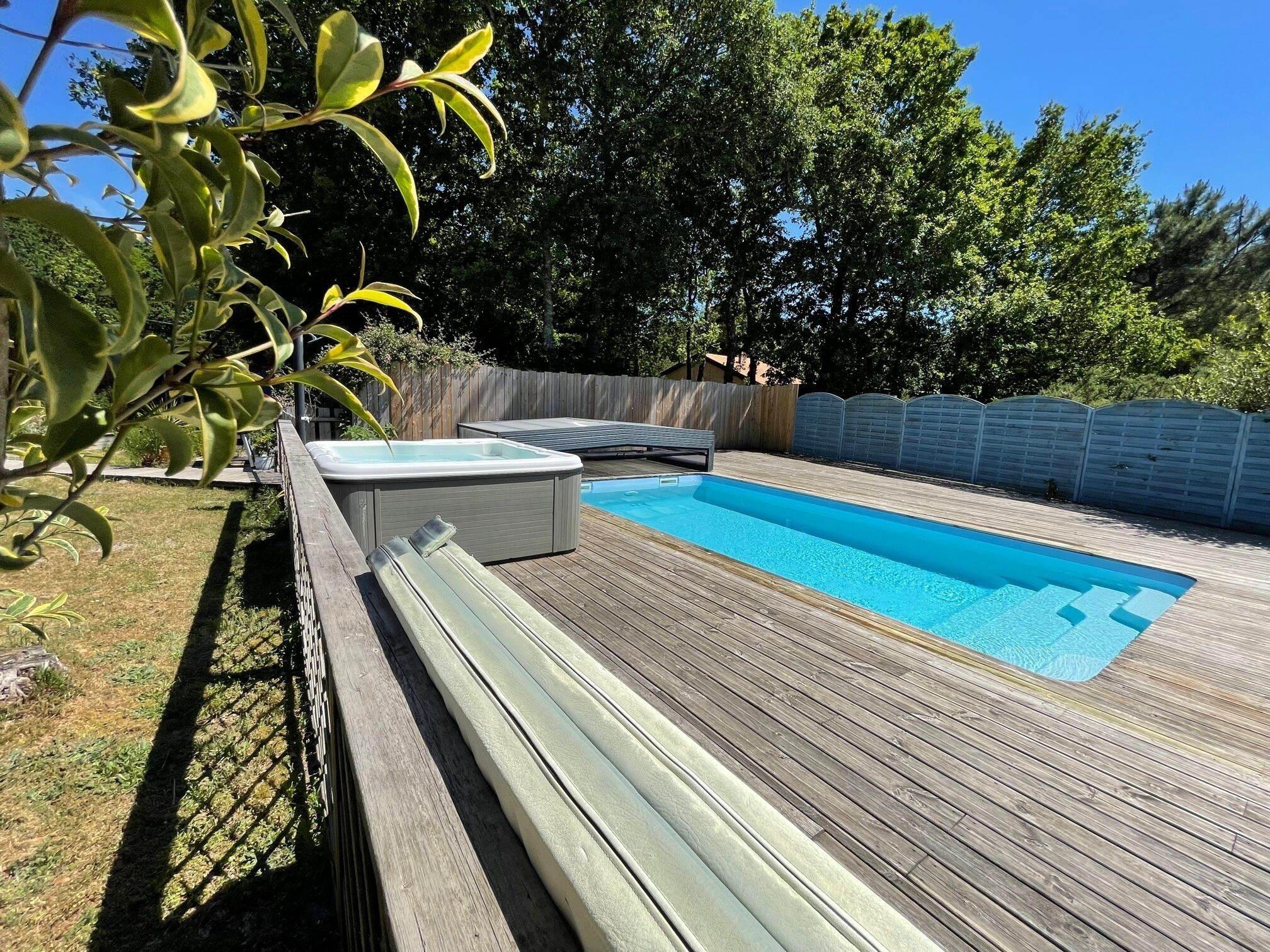 A rectangular swimming pool with a wooden deck, surrounded by a wooden fence and trees.