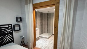 Un dormitorio con cama, mesita de noche, espejo y puerta de madera que da a un baño con ducha.