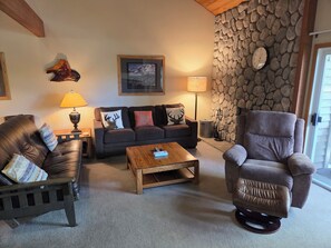 A cozy living room with a stone wall, a sofa, a recliner, and a coffee table. - Snowcreek Resort Vacation Rental 596 (Mammoth Lakes)
