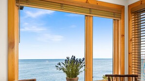 Una finestra con telaio in legno che si affaccia sul mare, un tavolo rotondo in legno con una pianta in vaso e due sedie di legno.