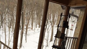 A cozy cabin interior with a large window offering a view of a snowy forest. There is a wooden table with books and a chair. - Cozy Cabin in The Driftless (Cross Plains)