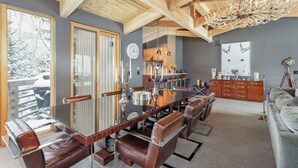 A spacious dining area with a long wooden table and leather chairs, a kitchen in the background, and a large window with a view of snow-covered trees.