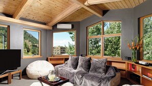 A modern living room with a grey sofa, a white pouf, and a wooden coffee table. Large windows offer views of a mountainous landscape.