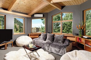 A modern living room with a grey sofa, a white pouf, and a wooden coffee table. Large windows offer views of a mountainous landscape.