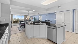 A modern kitchen with white cabinets, a black countertop, and a built-in dishwasher. There is a dining area with a table and chairs visible through an open door. - Yacht Club Villas #3-502 Wonderful Waterway Views! (North Myrtle Beach)