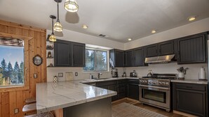 A modern kitchen with dark cabinets, a large island, stainless steel appliances, and a view of trees through the window. - Goetzl by Hauserman Rental Group (Tahoma)
