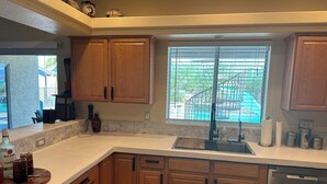 A kitchen with wooden cabinets, a stainless steel sink, and a window with a view of a pool and greenery. - Spacious Santa Fe 4b/4b Pool and Spa home centrally located (Lake Havasu City)