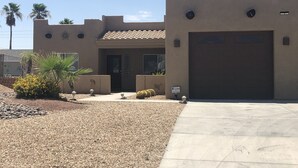 A building with a beige facade, a tiled roof, and a garage. There are palm trees and a small garden with yellow flowers. - Spacious Santa Fe 4b/4b Pool and Spa home centrally located (Lake Havasu City)