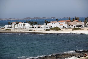 Complejo de edificios costeros con fachadas blancas y techos de tejas rojas, ubicado cerca de una playa de arena y el mar.