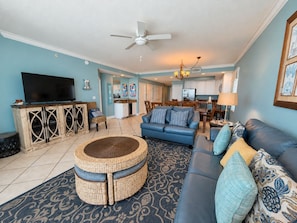 A living room with a blue sofa, a round wicker ottoman, a flat-screen TV, and a ceiling fan.