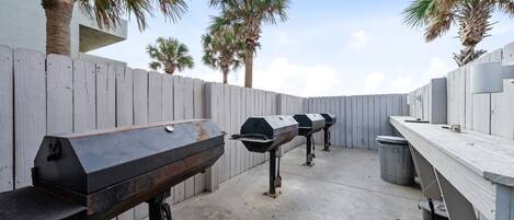 Un espace barbecue extérieur doté de plusieurs grils, d’une clôture en bois et de palmiers.