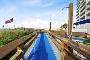 A wooden boardwalk leading to a beach with an American flag, a blue pathway, and a building with balconies. - Modern Beachfront Condo w/ Corner Views & Pool | Sleeps 10 (Gulf Shores)